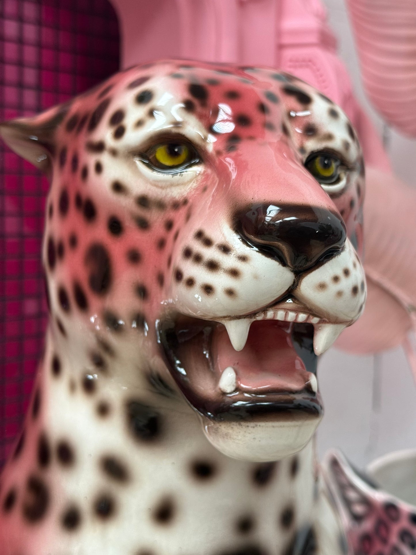 A close-up of a glossy ceramic leopard figurine with black spots on a pink and white body, yellow eyes, and an open mouth showing teeth, set against a pink background.