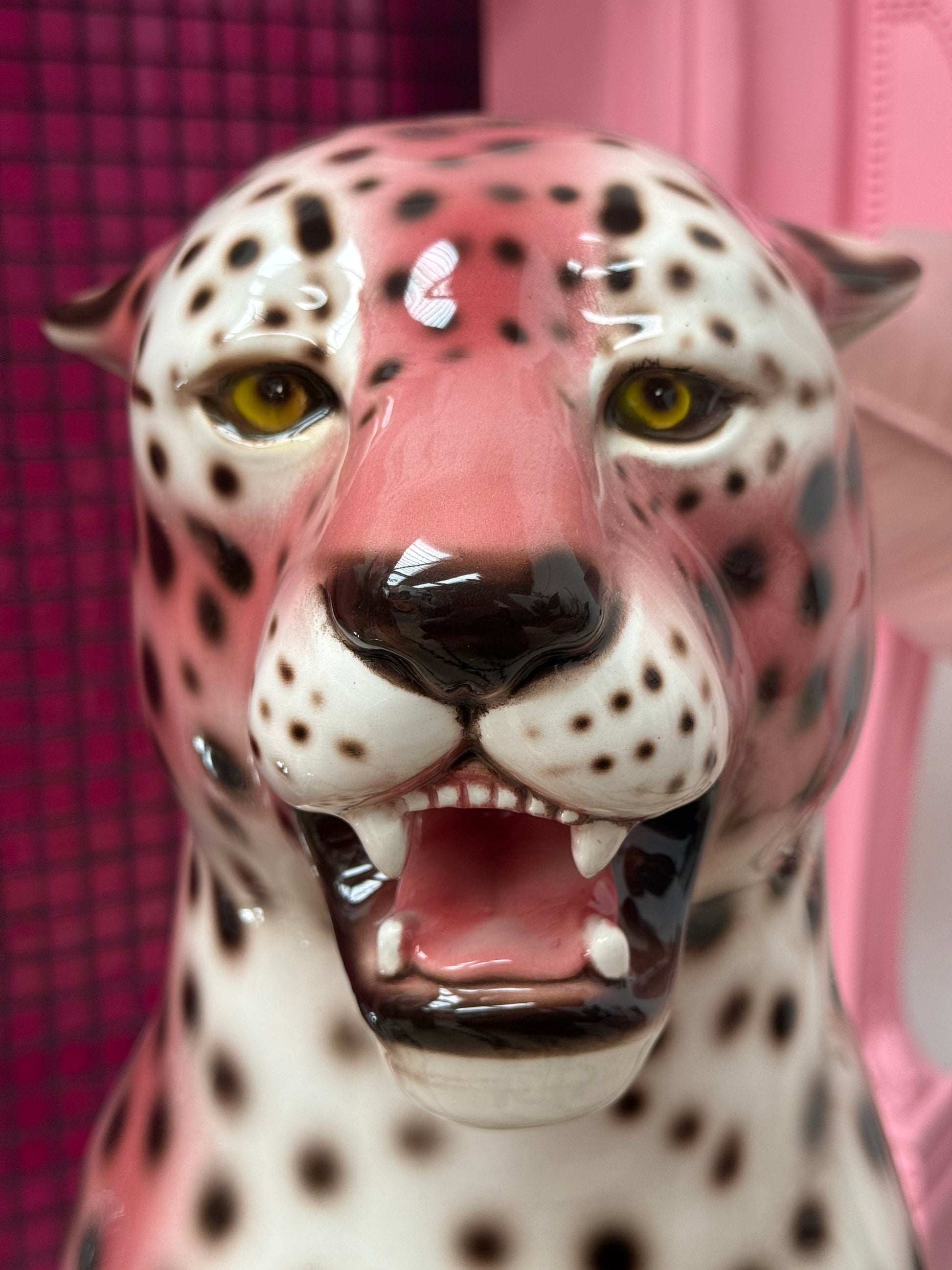 A close-up of a glossy ceramic leopard figurine with pink and white coloring, black spots, and an open mouth showing teeth, set against a pink background.