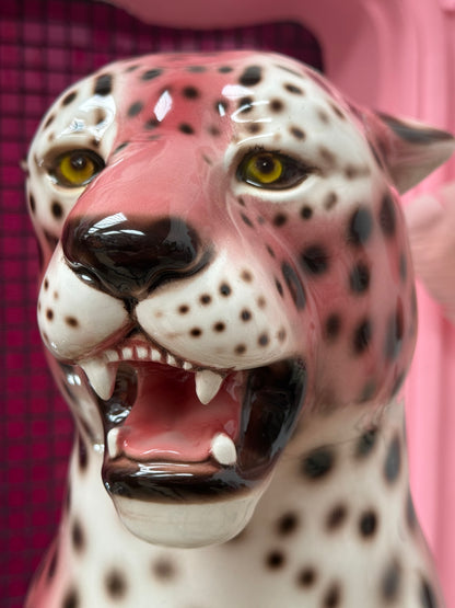 A close-up of a glossy ceramic leopard figurine with a pink and white spotted pattern, open mouth showing teeth, set against a pink background.