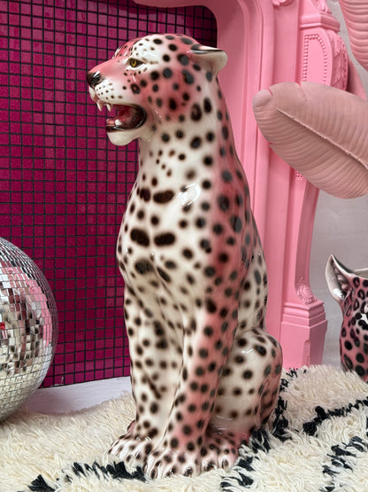 A glossy ceramic leopard figurine with a pink and cream spotted pattern, sitting on a black and white shag rug in a pink-themed room with a pink mirror, a disco ball, and a pink lip-shaped phone.