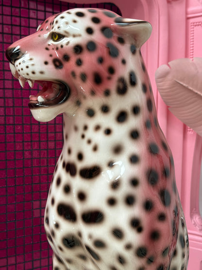 A glossy ceramic leopard figurine with a white body, black spots, and pink shading, shown close-up against a pink background.
